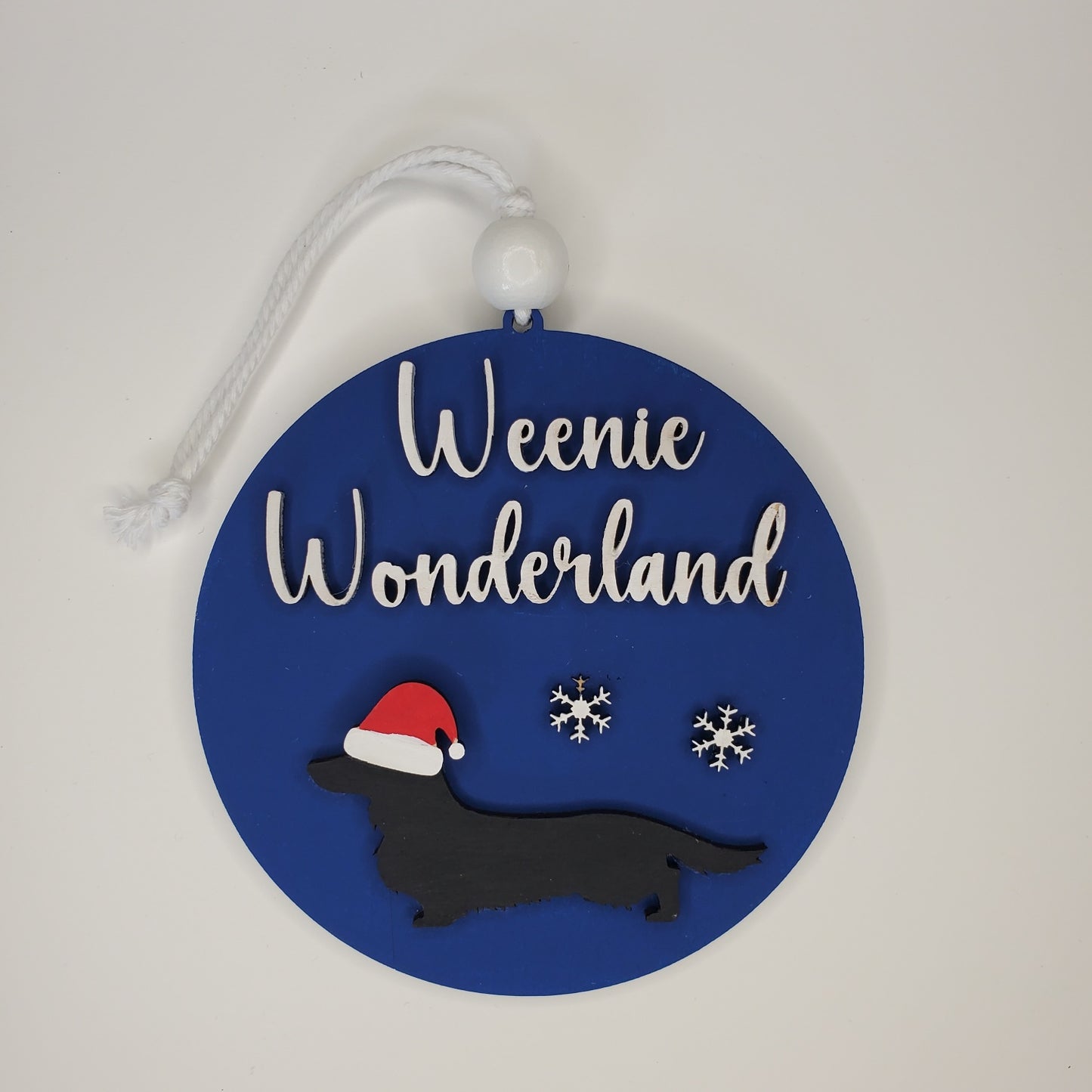 Decorative round ornament with 'Weenie Wonderland' text, dachshund silhouette, and snowflakes on a blue background. Wired, Smooth, and Long haired dogs available. Pick breed, dog color, and background color from available choices.