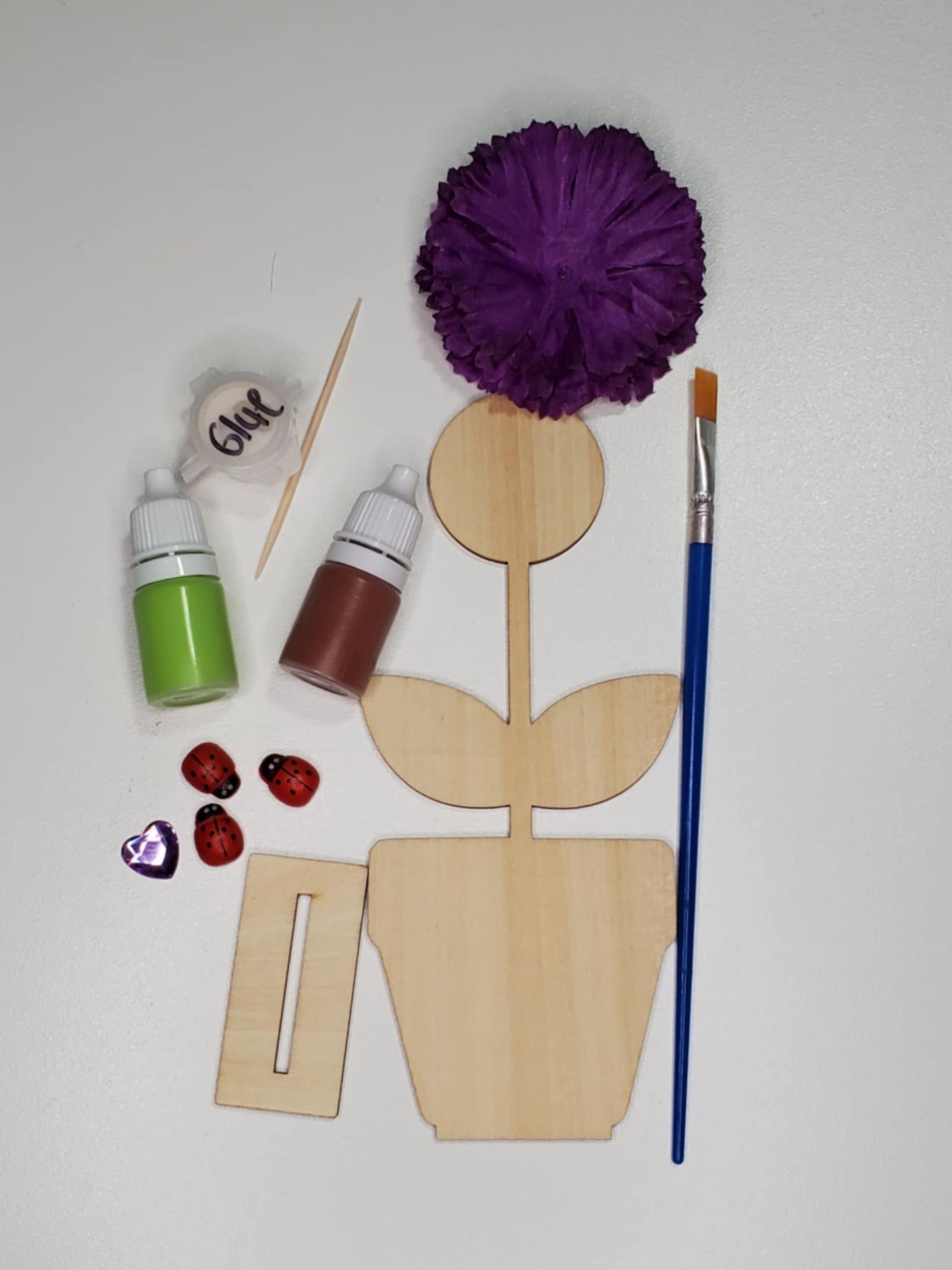 A laser cut wood flower pot and stem with a fake flower, lady bugs, paint, paintbrush, and glue to finish the project