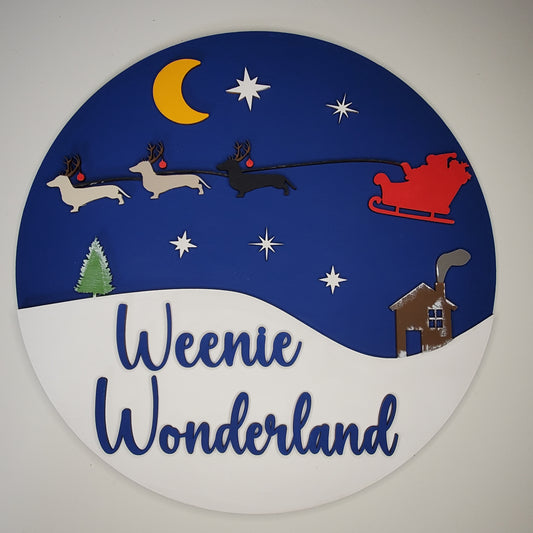 Wooden Door Round with dachshunds leading Santa's sleigh, a house, moon, and stars, and 'Weenie Wonderland' text on a blue and white background